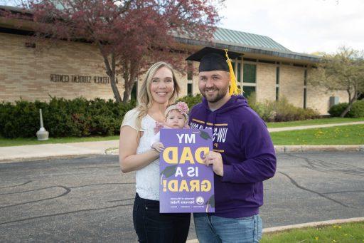 A male student in a grad cap posed with his wife and child outside to celebrate his masters degree..