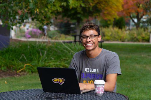 A student at a picnic table smiling and typing on a laptop computer with a coffee next to him.